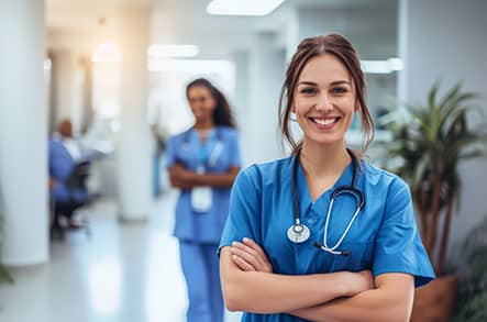 A smiling nurse looks at the camera, with another nurse standing in the distance behind her.